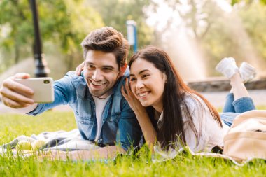 Akıllı telefondan selfie çeken ve parkta çimlerin üzerinde yatarken gülümseyen mutlu, çok kültürlü bir çift görüntüsü.