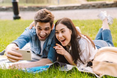 Mutlu, çok kültürlü bir çiftin selfie çekip parkta çimlerin üzerinde uzanırken akıllı telefonu işaret etmesi.