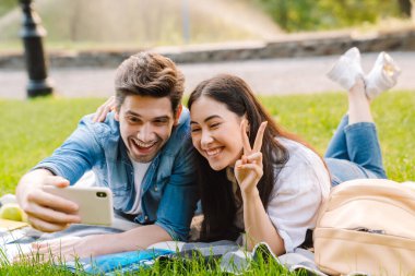 Akıllı telefondan selfie çeken ve parkta çimenlerde yatarken barış işareti yapan mutlu, çok kültürlü bir çiftin görüntüsü.