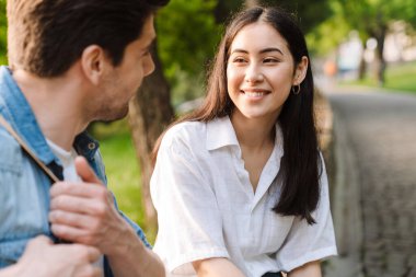 Parkta bankta otururken birbirleriyle konuşan ve gülümseyen neşeli, çok kültürlü çiftin görüntüsü