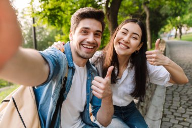 Neşeli, çok kültürlü bir çiftin selfie çekerken ve parkta bankta otururken başparmaklarını gösterirken görüntüsü