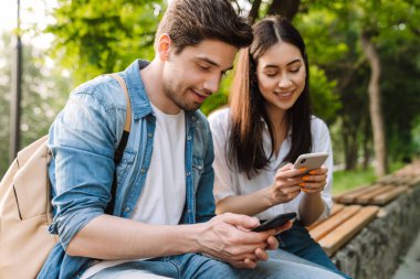 Cep telefonu kullanan ve parkta bankta otururken gülümseyen çok kültürlü bir çift görüntüsü