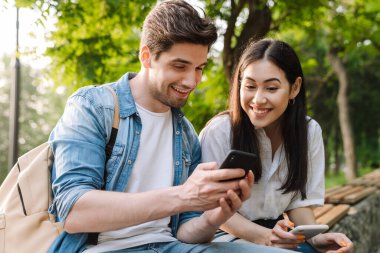 Cep telefonu kullanan ve parkta bankta otururken gülümseyen çok kültürlü bir çift görüntüsü