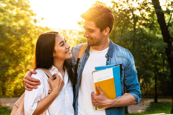 Image of cheerful multicultural student couple hugging and smiling while walking in green park