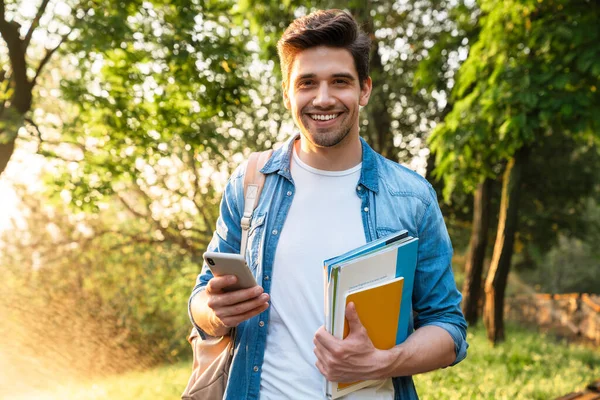 Photo of smiling unshaven student man with exercise books using smartphone while walking in green park