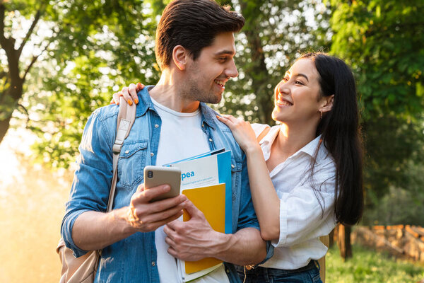 Image of cheerful multicultural student couple using mobile phone and smiling while walking in green park