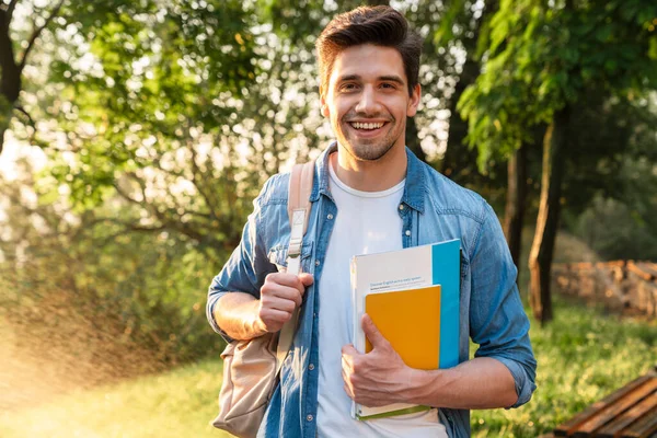 Photo of happy unshaven student man with exercise books smiling at camera while walking in green park