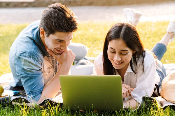 在公园里躺在草地上笑着使用笔记本电脑的快乐的多文化夫妇的形象 — 图库照片