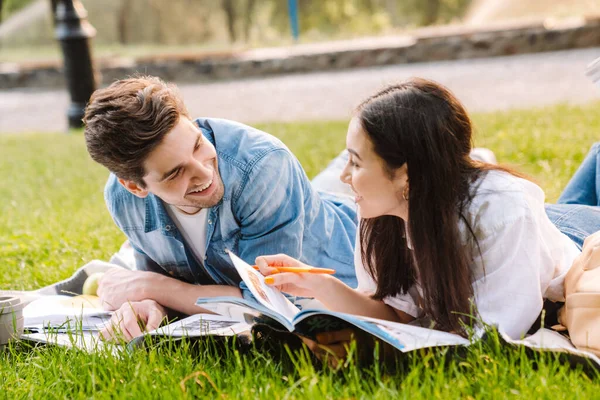 公園で草の上に横になりながら宿題や笑顔をする楽しい多文化学生カップルのイメージ — ストック写真