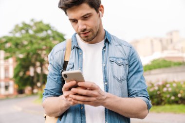 Kablosuz kulaklıklı, cep telefonlu, sırt çantalı, bulvarda yürürken çekilmiş bir fotoğraf.