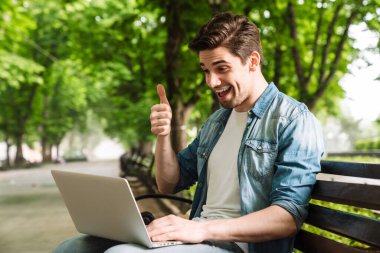 Green Park 'ta bankta otururken dizüstü bilgisayar kullanan heyecanlı genç adamın fotoğrafı.