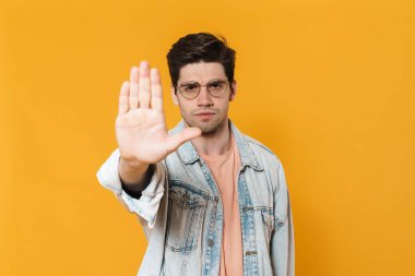 Gözlüklü genç adamın sarı arka planda izole edilmiş dur hareketi gösteren fotoğrafı.