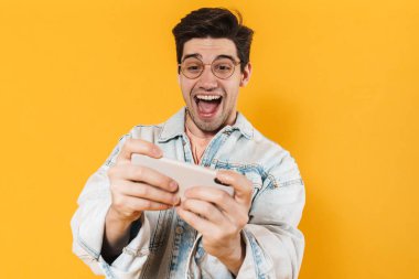 Sarı arka planda izole edilmiş cep telefonuyla online oyun oynayan heyecanlı genç adamın fotoğrafı