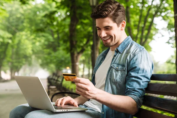 Foto Jovem Feliz Usando Laptop Segurando Cartão Crédito Enquanto Sentado — Fotografia de Stock