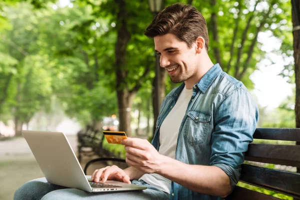 緑の公園のベンチに座っている間 ノートパソコンとクレジットカードを持って幸せな若い男の写真 — ストック写真