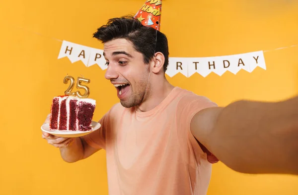 Photo of excited caucasian man in party cone taking selfie photo with birthday cake isolated over yellow background