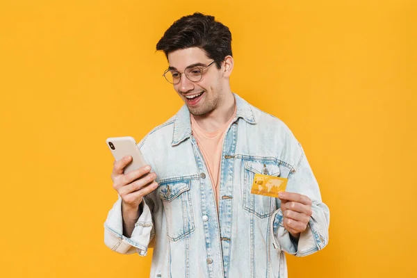 写真はクレジットカードを保持し 黄色の背景に隔離された携帯電話を使用して喜んで若い男の — ストック写真