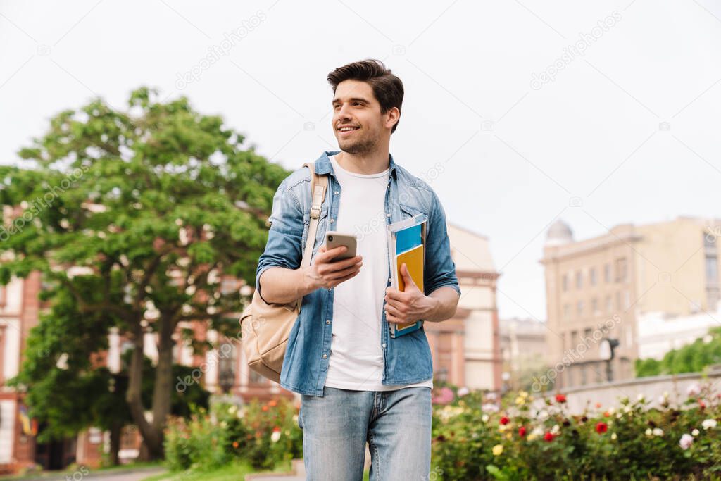 Photo of happy handsome man smiling and using cellphone while walking with backpack on boulevard