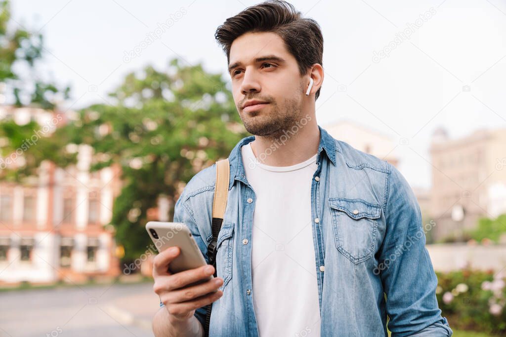 Photo of focused man with wireless earphone using cellphone while walking with backpack on boulevard