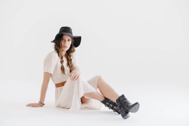 Şapkalı, esmer, baştan çıkarıcı bir kadının beyaz arka planda izole bir şekilde poz verdiği ve oturduğu fotoğraf.