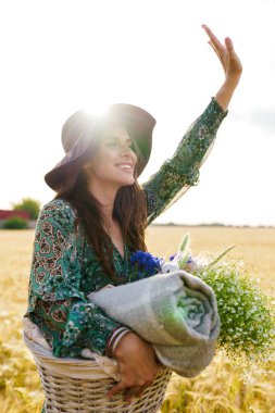 Buğday tarlasında piknikte sepetle yürürken şık şapkalı neşeli bir kadının fotoğrafı.