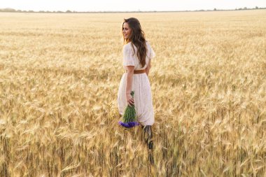 Güzel, mutlu, şık elbiseli bir kadının fotoğrafı buğday tarlasında çiçeklerle yürürken gülümsüyor.