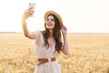Yazın buğday tarlasında yürürken cep telefonuyla konuşan güzel bir kadının fotoğrafı.