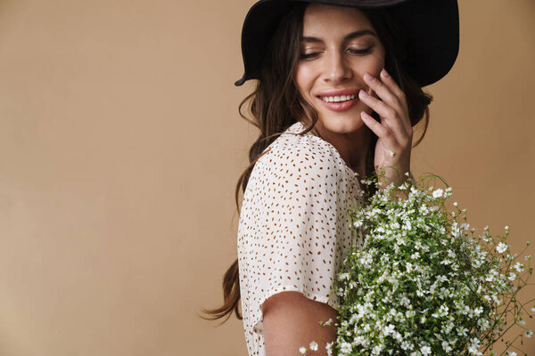 Photo of cheerful beautiful woman in hat smiling while posing with flowers isolated over beige background