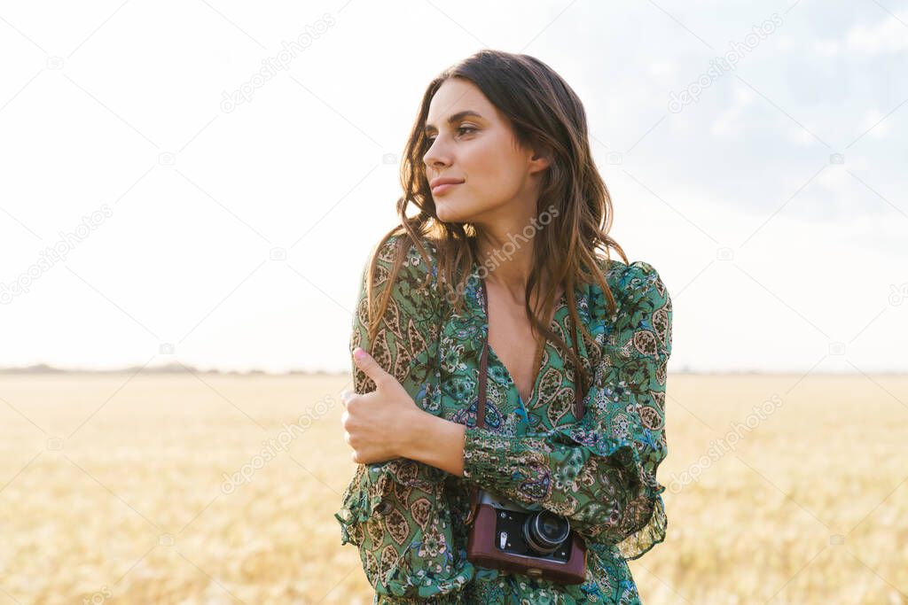 Photo of pleased cute woman in stylish dress looking aside while walking with retro camera on wheat field