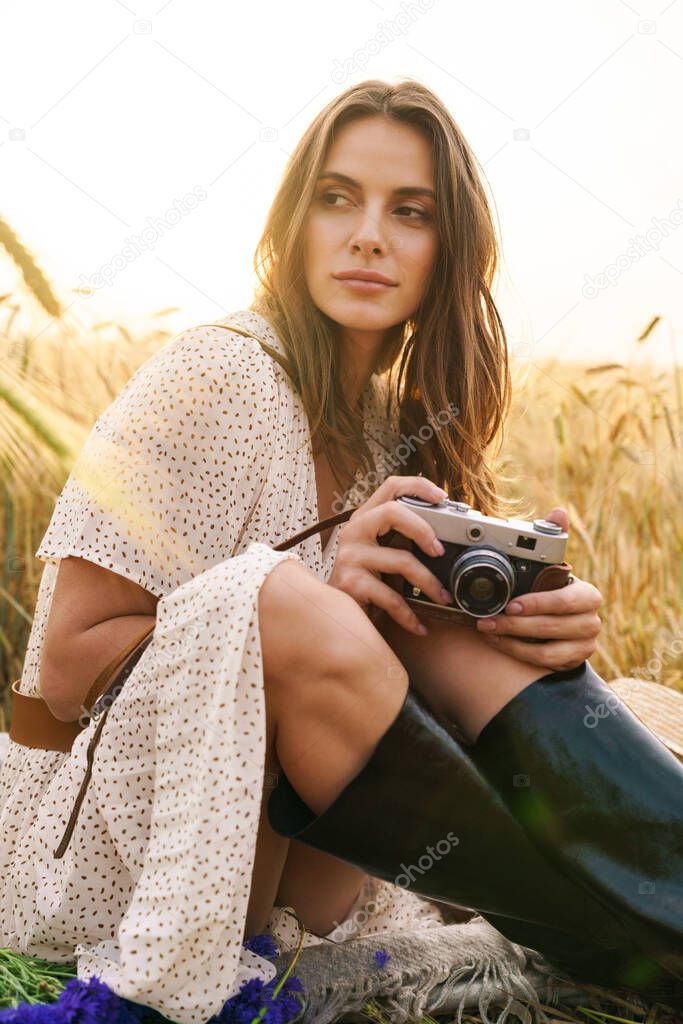 Photo of charming young woman in stylish dress holding retro camera while sitting on wheat field