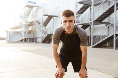 Kulaklıklı, traşsız, beyaz bir sporcunun şehir dışında egzersiz yaparken görüntüsü.