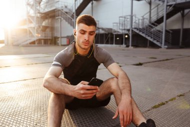 Kafkasyalı genç bir sporcunun dışarıda otururken cep telefonu ve kulaklık kullanması.