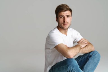 Gri arkaplanda izole bir şekilde otururken kameraya bakan kızıl saçlı, erkeksi bir adamın portresi.