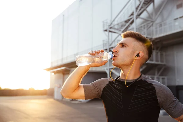 屋外の都市部で仕事をしながら水を飲むイヤホンの若いスポーツマンのイメージ — ストック写真