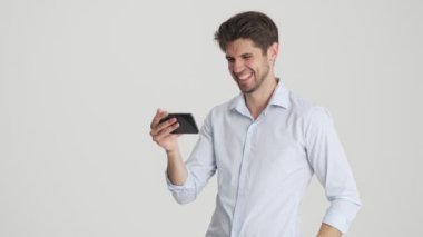 Gülen bir adam, beyaz arka planda yatay olarak duran akıllı telefonuyla bir şey izliyor.