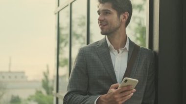 Gülümseyen yakışıklı işadamı binanın yanında dikilirken akıllı telefon kullanıyor.