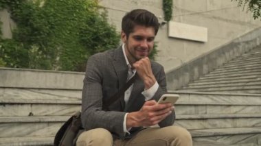 Gülümseyen yakışıklı işadamı, dışarıdaki bankta otururken akıllı telefon kullanıyor.