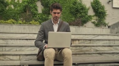 Sakin yakışıklı iş adamı dışarıda merdivenlerde otururken dizüstü bilgisayar kullanıyor.