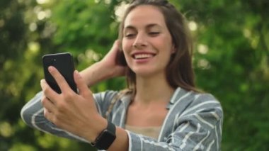 Güzel, çekici esmer kadın akıllı telefondan selfie çekiyor ve parkta saçını düzeltiyor.
