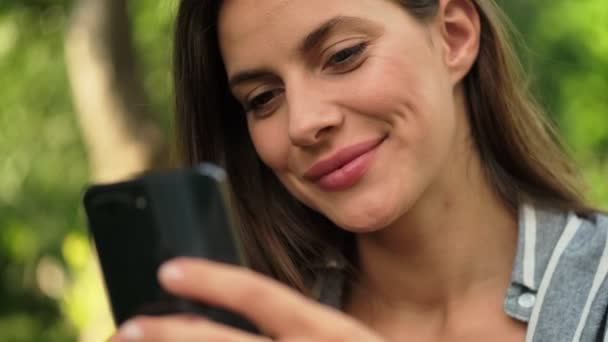 公園に座っている間にスマートフォンを使用して幸せな魅力的なブルネットの女性 — ストック動画