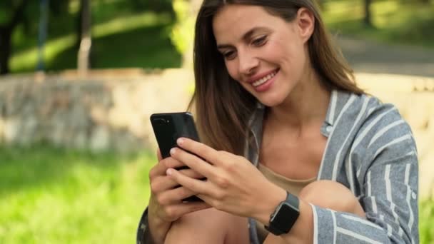 Uśmiechnięta Atrakcyjna Brunetka Korzystająca Smartfona Siedząc Parku — Wideo stockowe