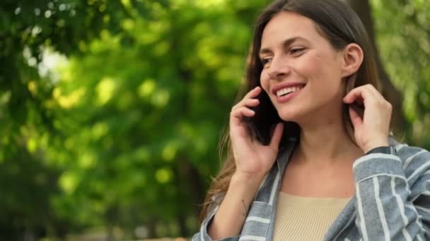 笑顔魅力的なブルネットの女性はスマートフォンで話し 公園で見て回る — ストック動画