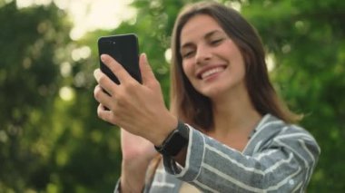 Neşeli, çekici esmer kadın akıllı telefondan video konuşmalarıyla konuşuyor ve parkta kameraya el sallıyor.