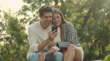 Parkta birlikte otururken akıllı telefon kullanan mutlu genç çift.