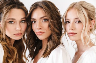 Beyaz duvarın üzerinden izole edilmiş, poz veren ve kameraya bakan beyaz, çekici üç kadının fotoğrafı.