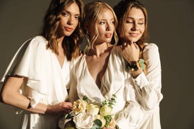 Gri duvarda saklanmış çiçeklerle poz veren beyaz elbiseli güzel gelininin fotoğrafı.