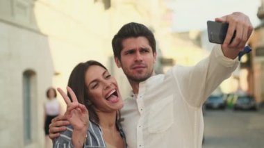 Neşeli, genç ve güzel bir çift sokakta yan yana dururken akıllı telefondan selfie çekiyor.