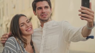 Tatmin olmuş genç bir çift sokakta yan yana dururken akıllı telefondan selfie çekiyor.