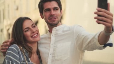 Gülümseyen genç ve güzel bir çift sokakta yan yana dururken akıllı telefondan selfie çekiyor.