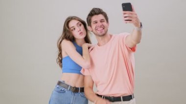 Mutlu, genç bir çift, stüdyodaki gri arka planda tek başına duran akıllı telefondan selfie çekiyor.